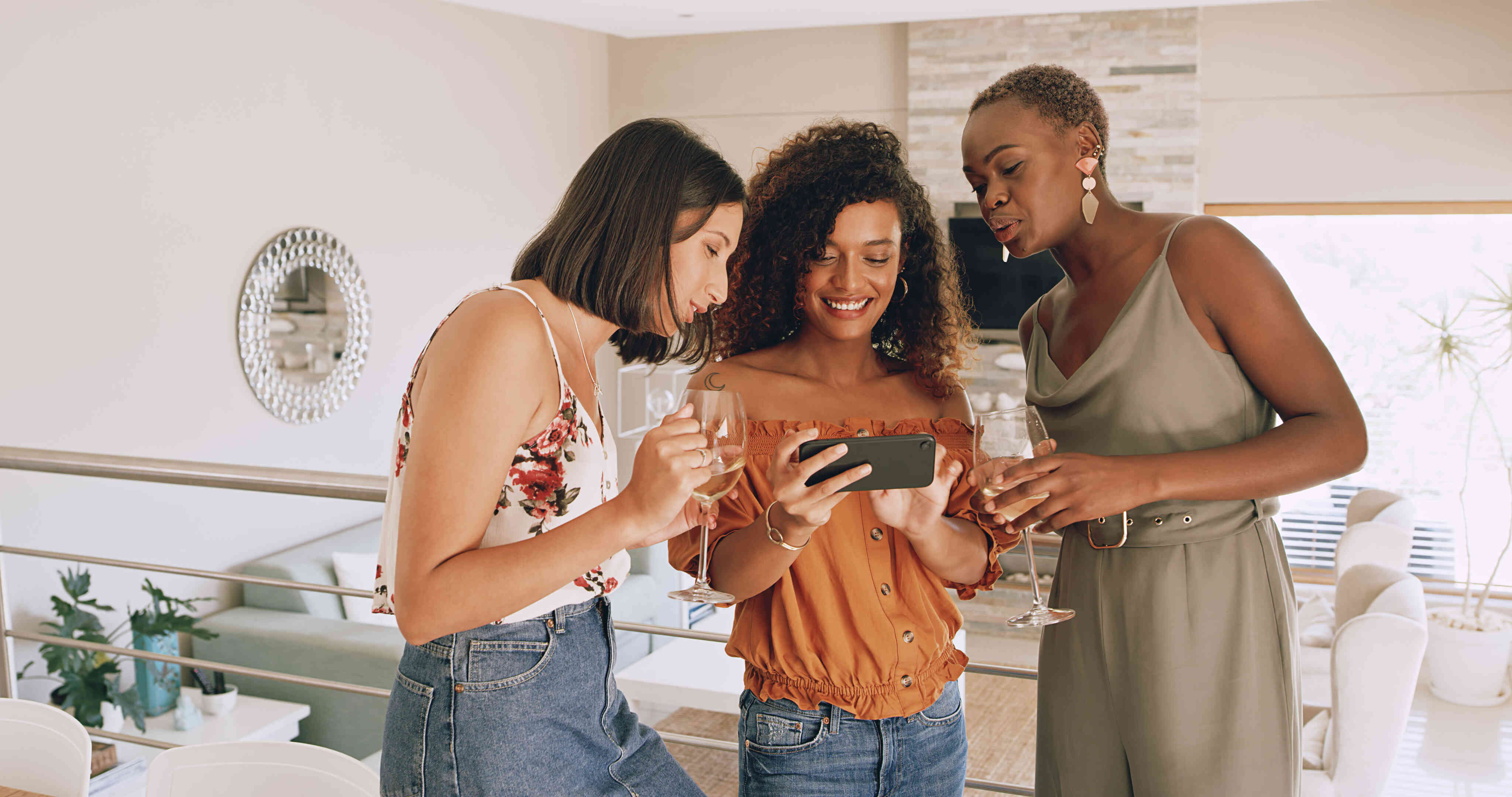 Three women stand close together in a home while all looking at the phone in one woman's hands.